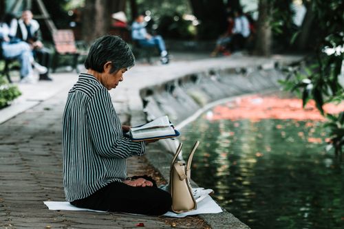 Free Elderly woman enjoying a peaceful reading moment by a pond in a serene park setting. Stock Photo