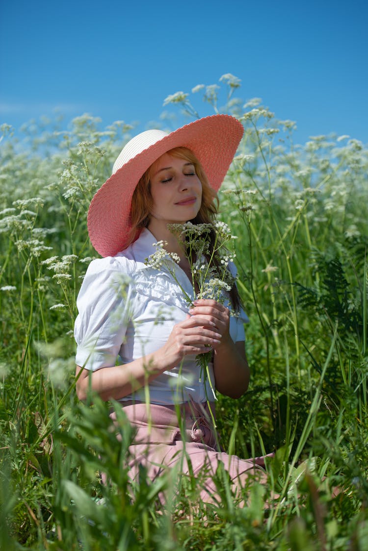 Woman In Pink Straw Hat And White Blouse Sitting Holding A Bunch Of Wildflowers