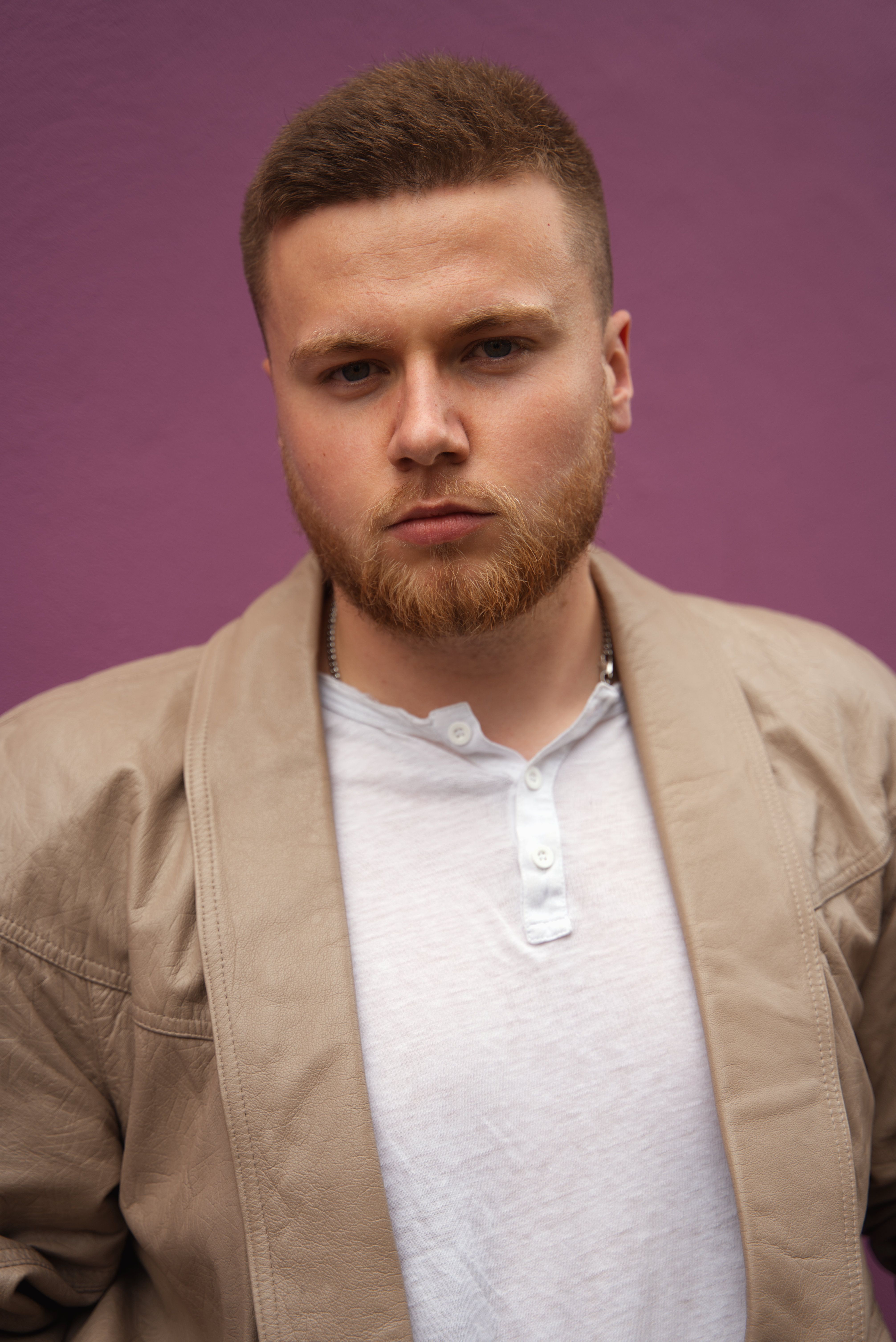 Casual portrait of a stylish bearded man against a purple background, showcasing modern fashion.
