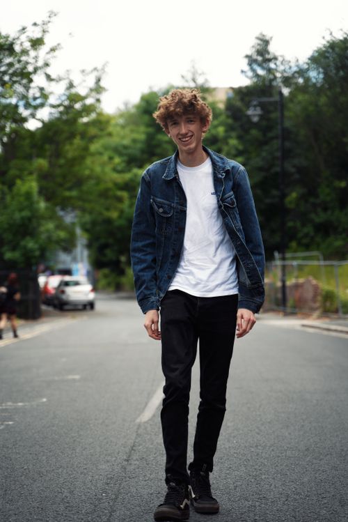 Free Smiling young man with curly hair walks confidently in a jean jacket on a city road. Stock Photo