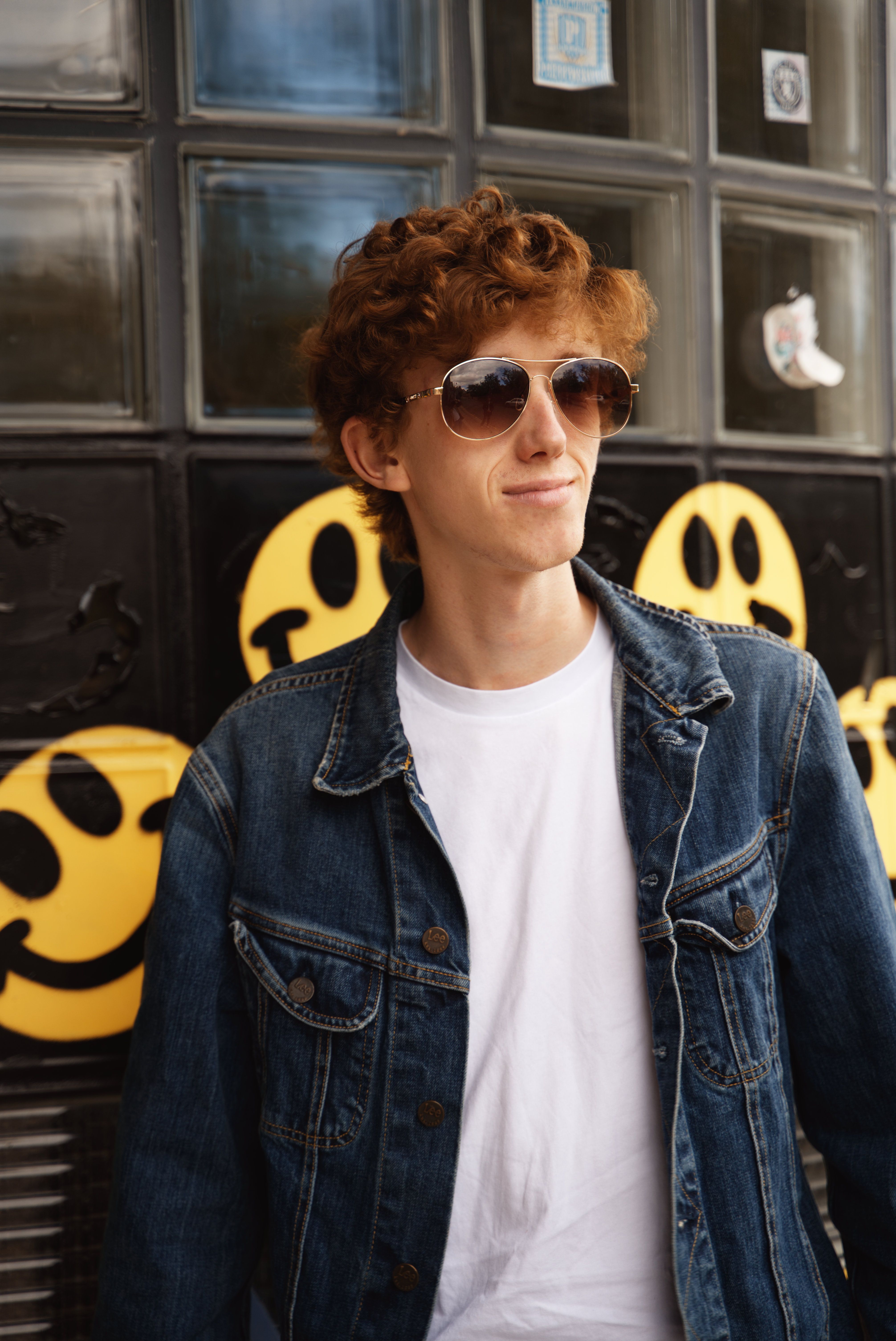 Stylish redhead teenager wearing sunglasses and a denim jacket, posing against a graffiti wall with smiley faces.