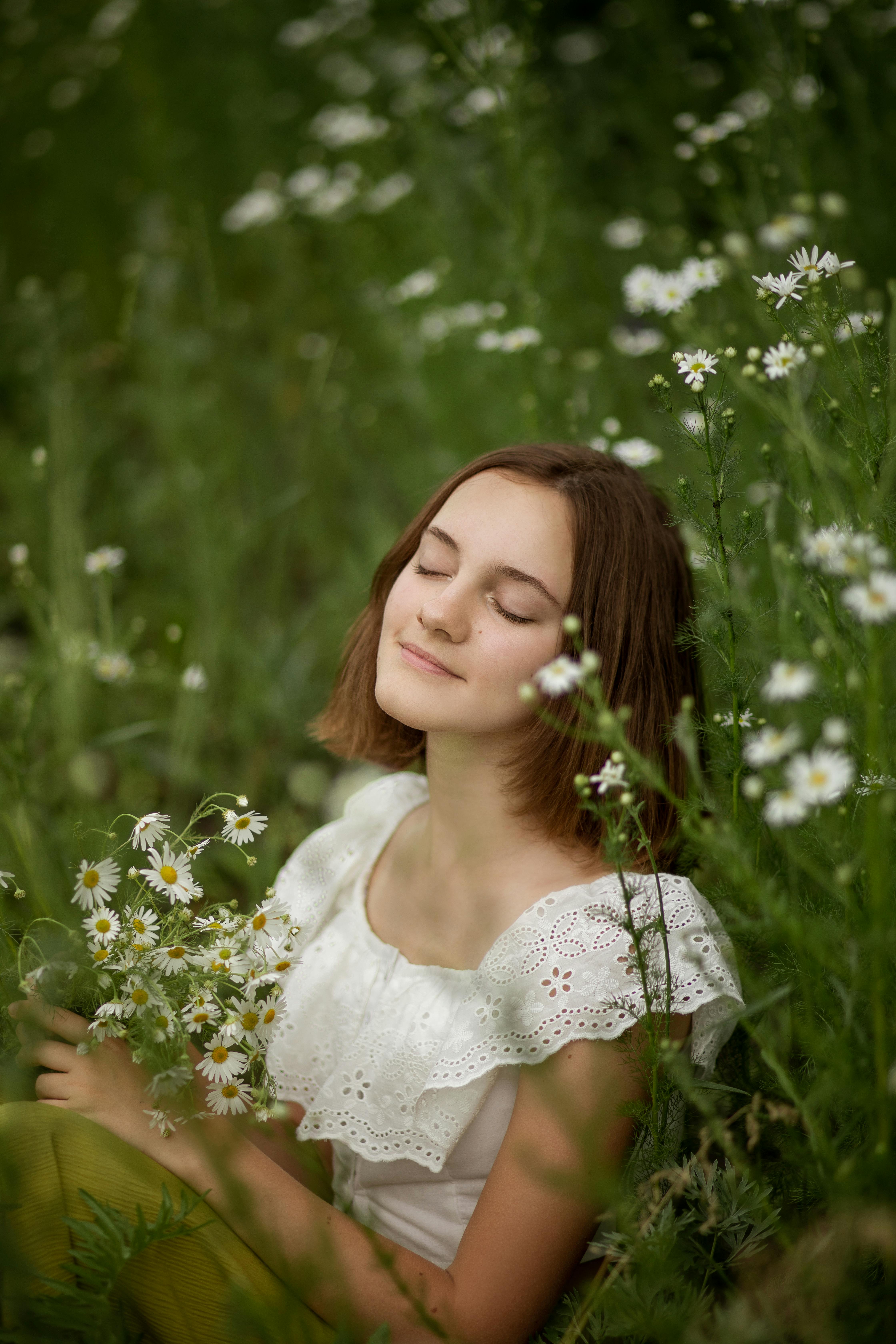 A woman with closed eyes peacefully sits in a meadow surrounded by wildflowers.