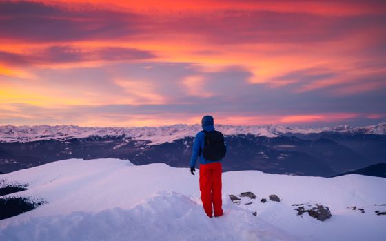 A lone hiker admires the breathtaking alpine sunset hues from a snowy peak in the Italian Alps.