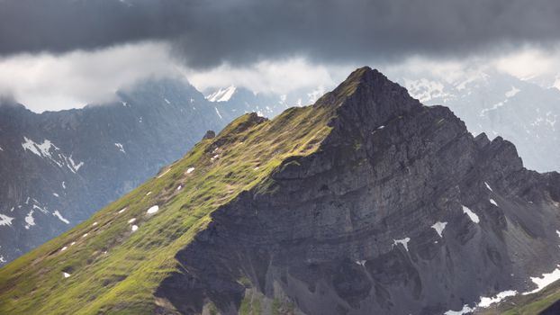 Dramatic mountain landscape with rocky peaks, green slopes, and swirling clouds.
