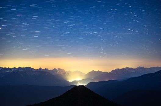 Long exposure of starry night sky casting light over rugged Alpine mountain range.