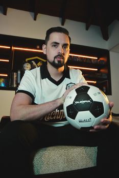 Man posing indoors holding a soccer ball while wearing a sports uniform, creating an athletic ambiance.