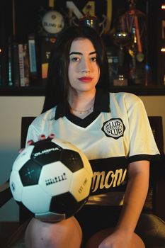 A woman seated indoors holding a soccer ball, wearing a sports club shirt.