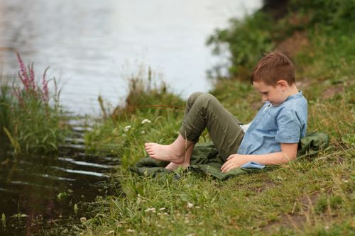 Free A young boy sitting by a tranquil stream, surrounded by green grass and wildflowers. Stock Photo