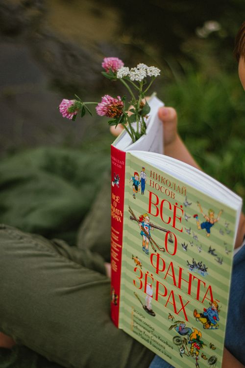 Free A child holding a Russian book with flowers, enjoying a peaceful outdoor reading session. Stock Photo