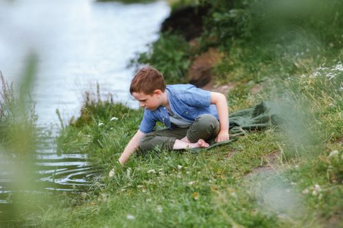 Free A young boy with brown hair sits by a rural riverbank, gazing at the water on a warm summer day. Stock Photo