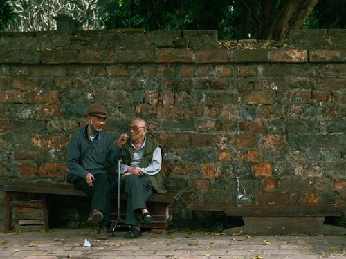 Free Two elderly men sit and chat on a bench against a rustic stone wall in Hanoi, Vietnam. Stock Photo