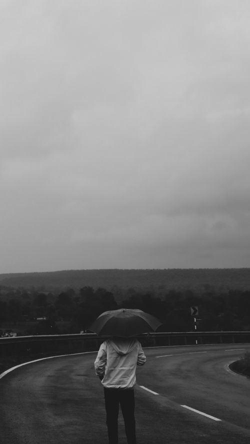 Free A lone man stands with an umbrella on a deserted rainy road, evoking a sense of solitude. Stock Photo
