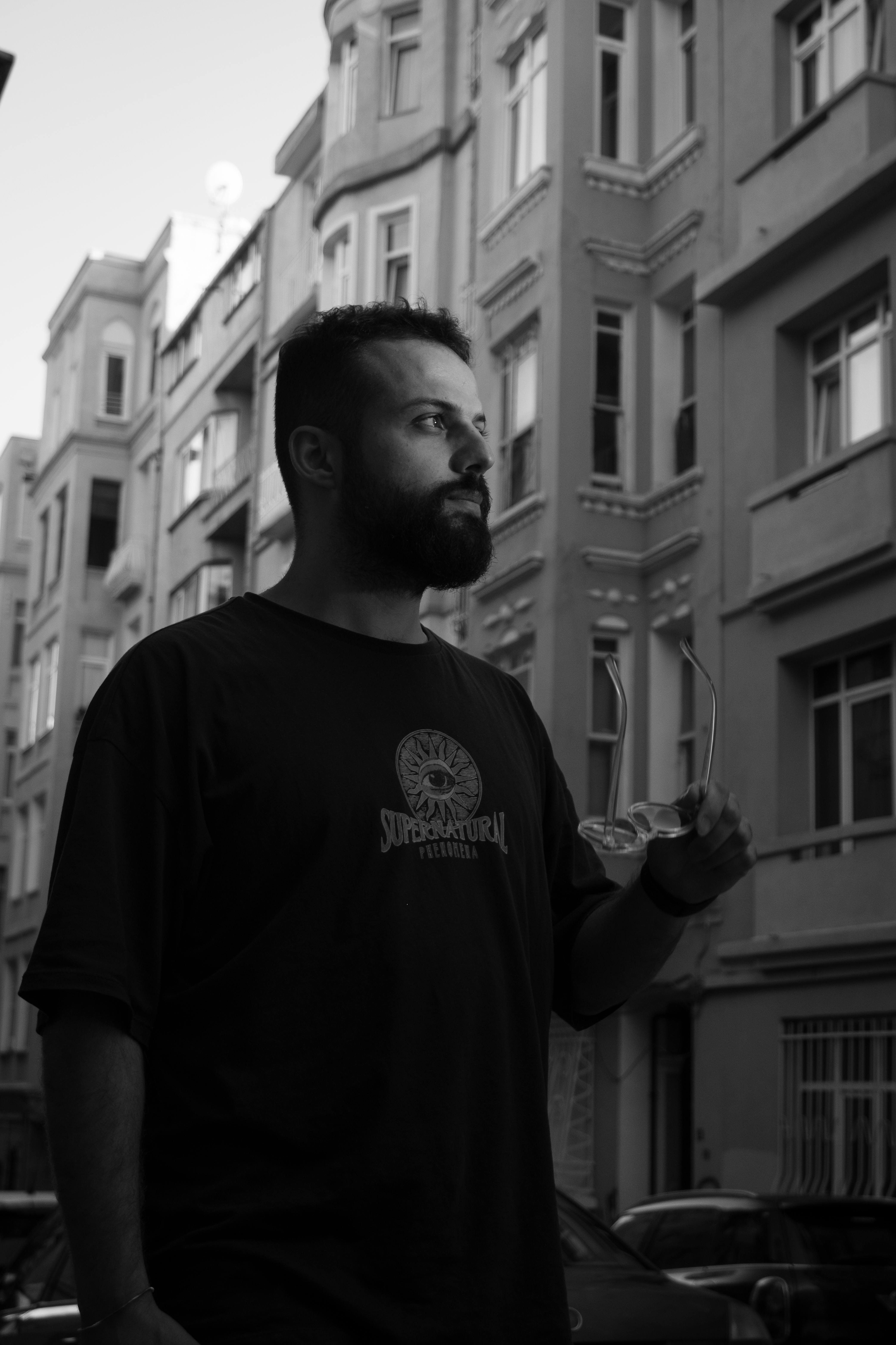 A black and white portrait of a man with a beard and eyeglasses in an urban setting.