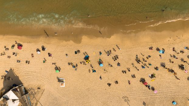 Drone aerial view of a sunlit beach with people lounging and playing by the shore in Rio de Janeiro.
