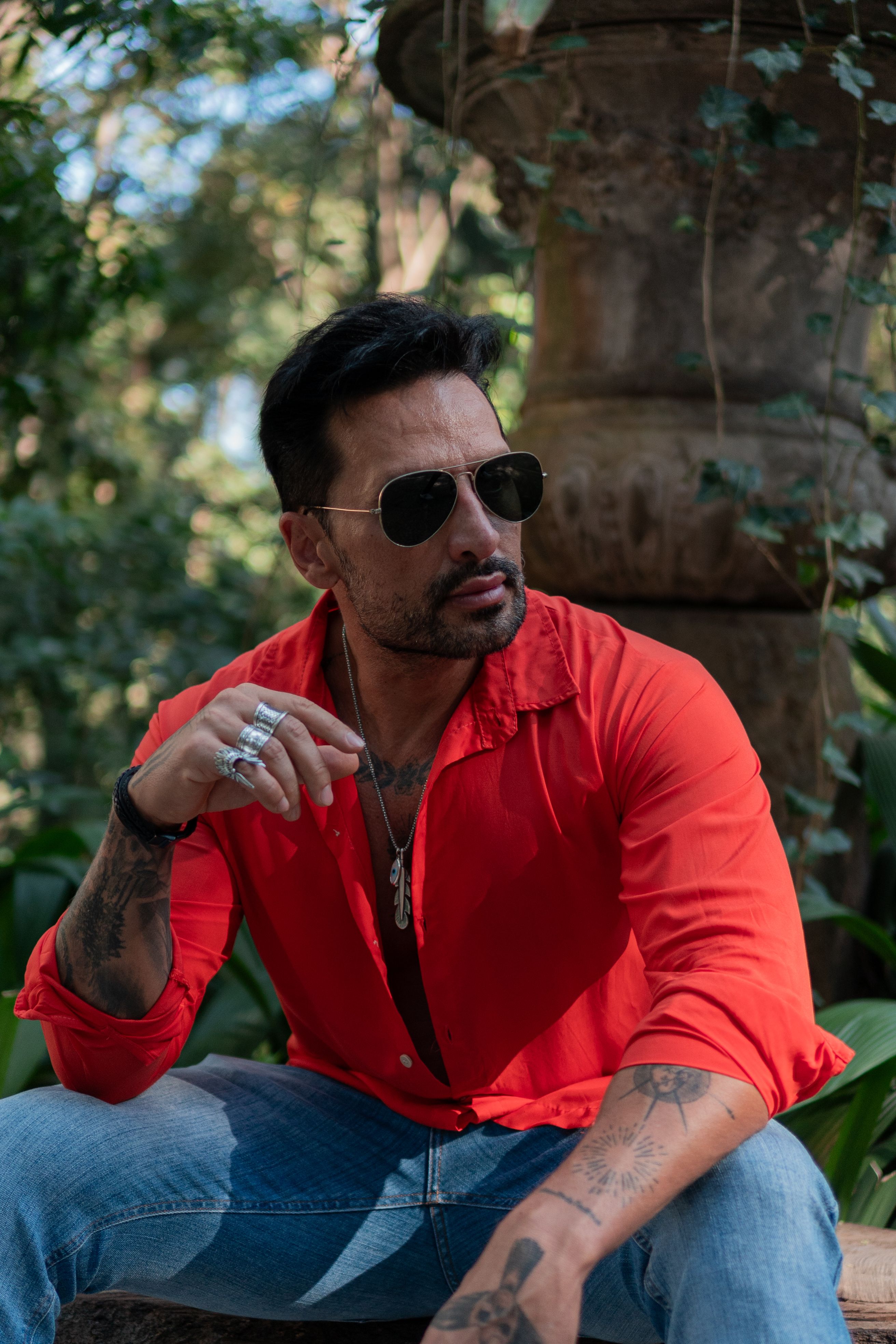 Homem fashion com camisa vermelha sentado ao ar livre, usando óculos de sol e tatuagens, exalando estilo.