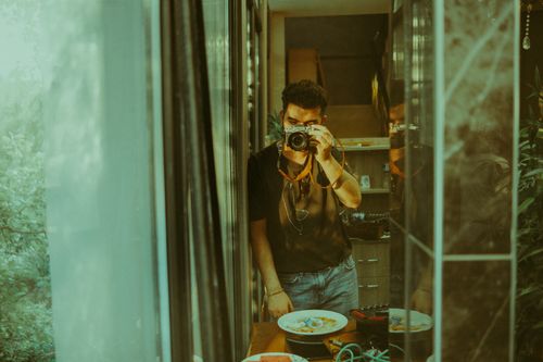 Free A man holding a vintage camera capturing his reflection in a mirror indoors, with a warm sepia tone. Stock Photo