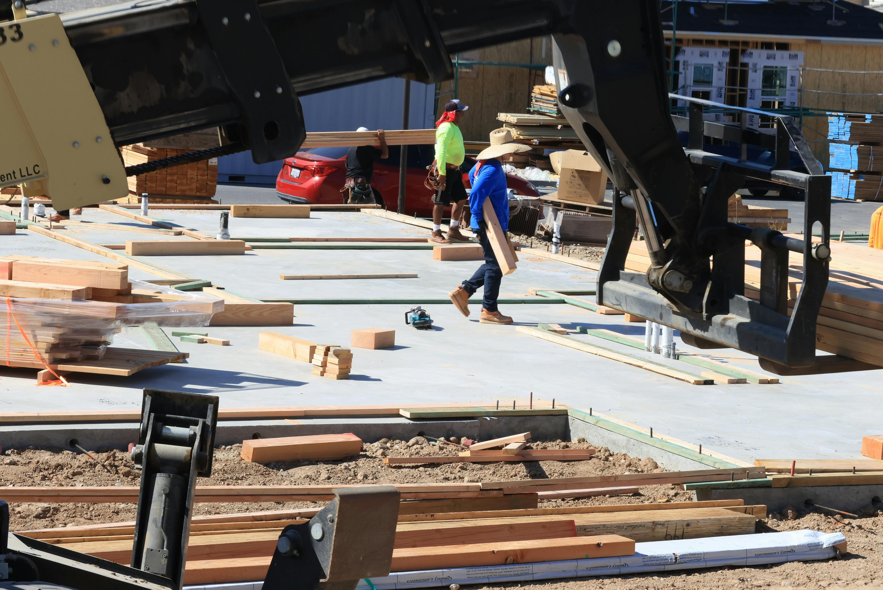 Construction workers on a building site, one carrying wood, with an excavator in the foreground.