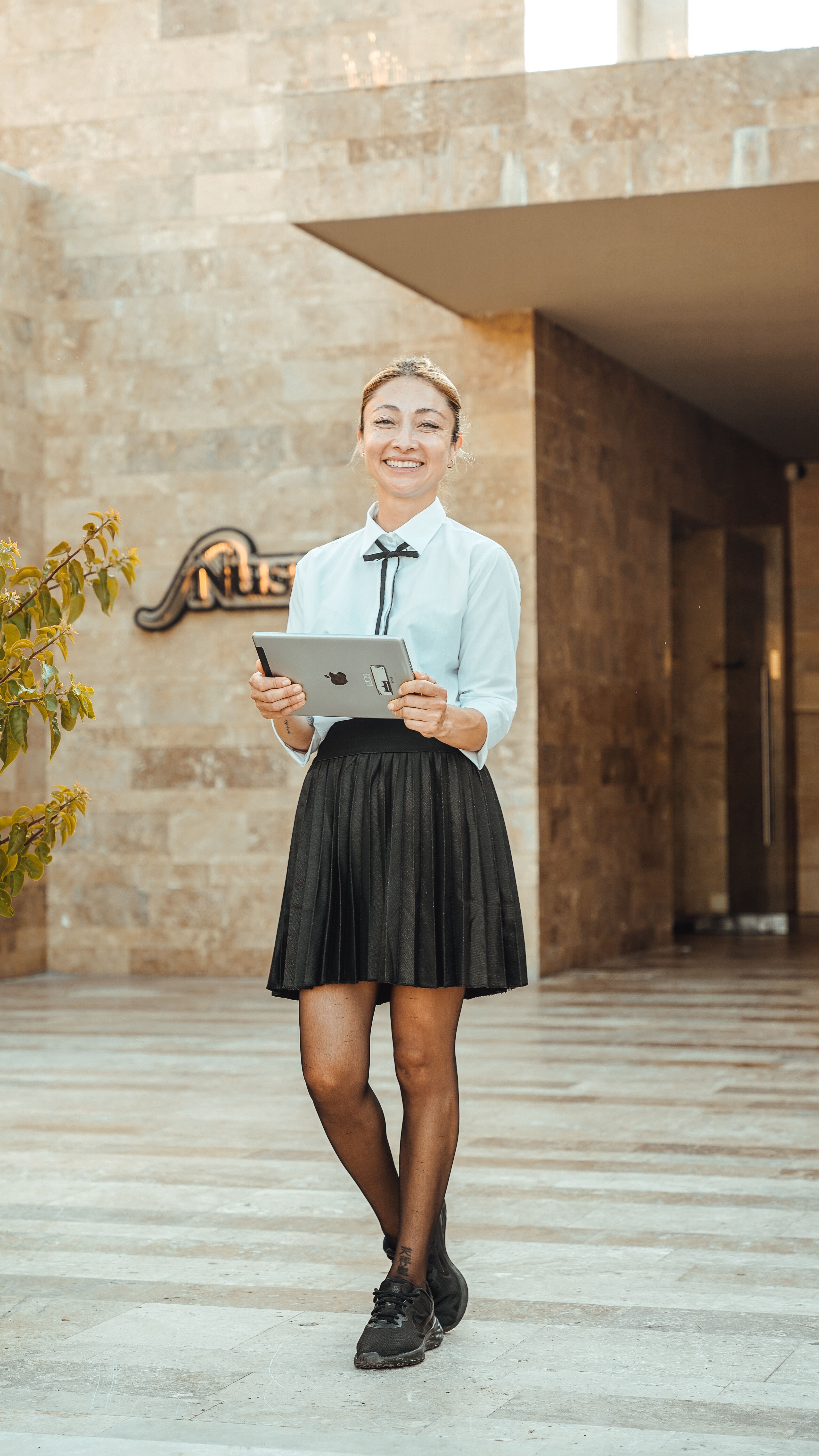 A young professional woman stands outdoors holding a tablet, smiling confidently.