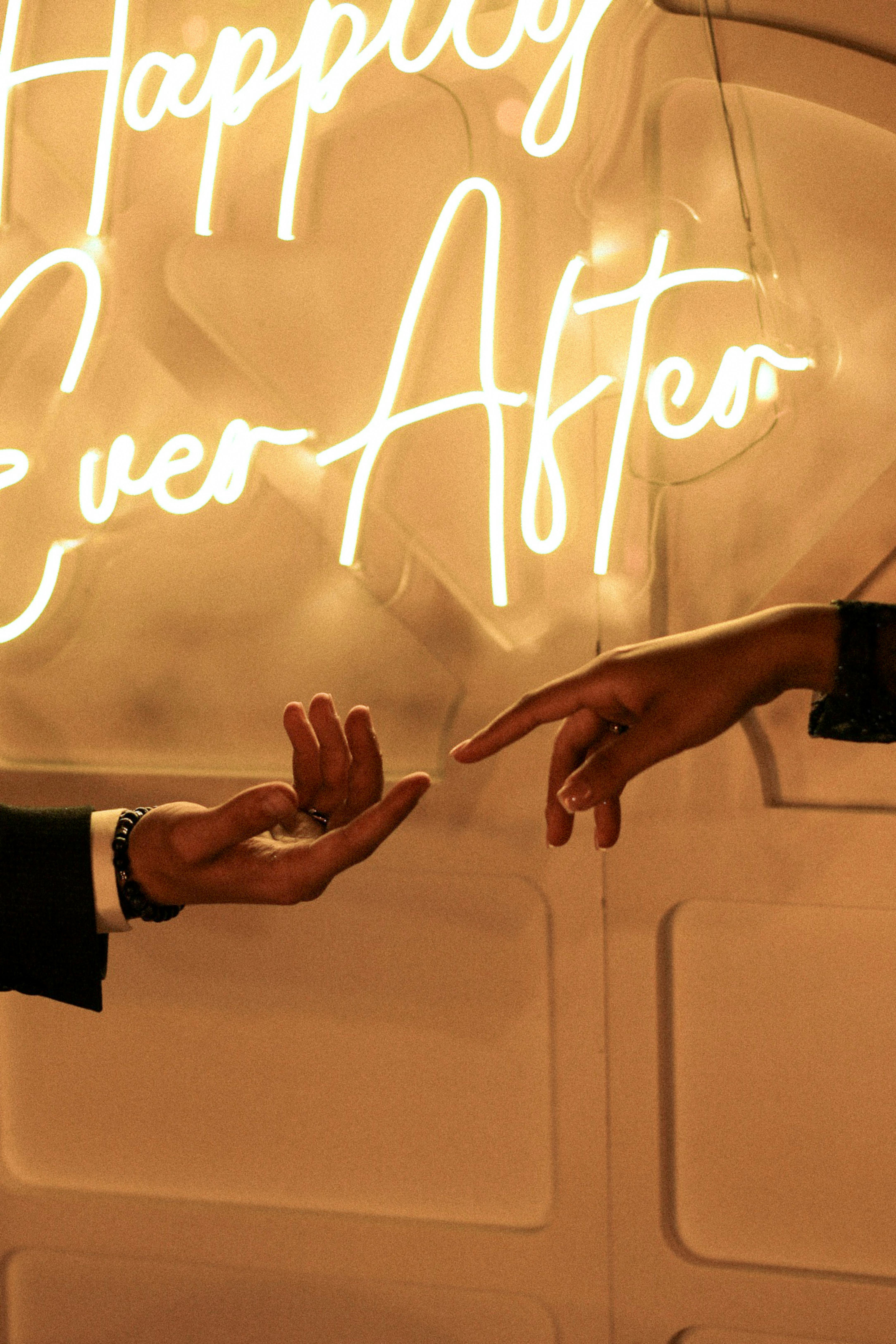 Hands reaching beneath a 'Happily Ever After' neon sign, evoking romance and connection.