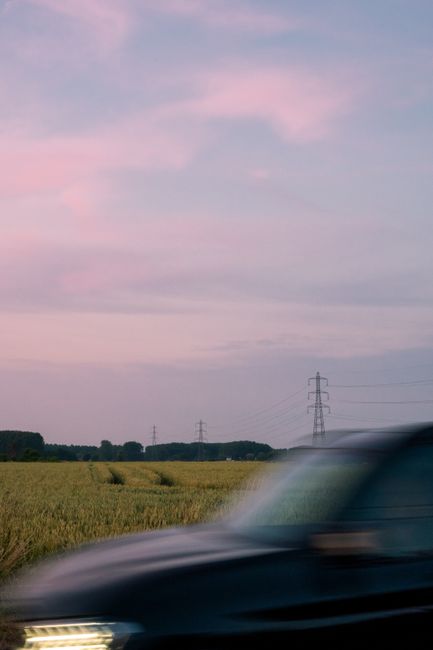A blurred car drives down a rural road during sunset, with fields and power lines in the background.