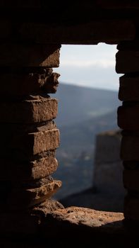 A closeup of a rustic stone wall with a window opening, revealing a distant mountain view.