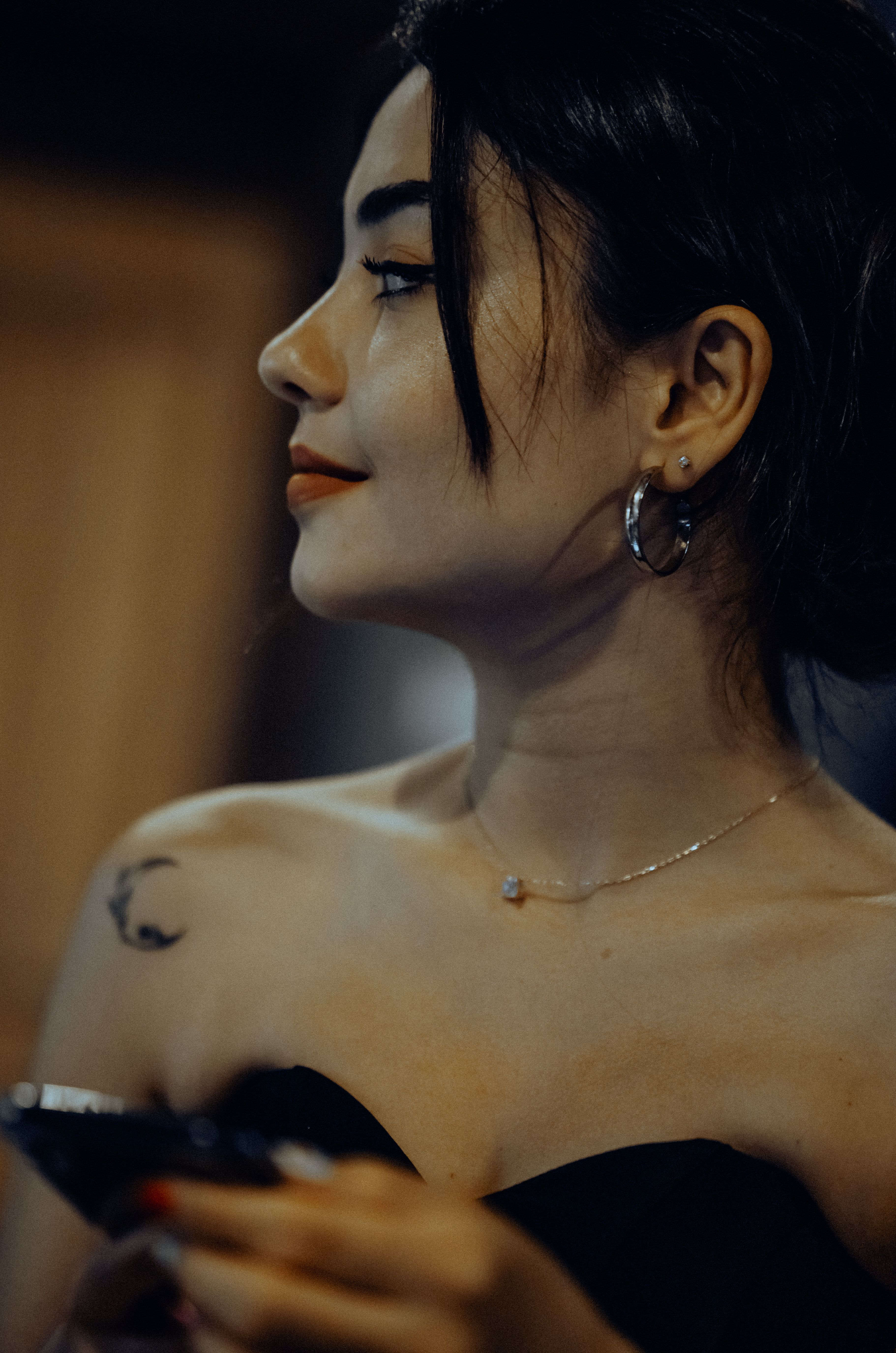 Profile of a woman with black hair, tattoo, and necklace in a dimly lit setting.