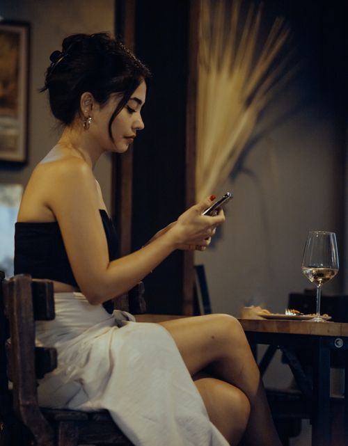 Free A fashionable woman sitting indoors, using a smartphone with a glass of wine on the table. Stock Photo