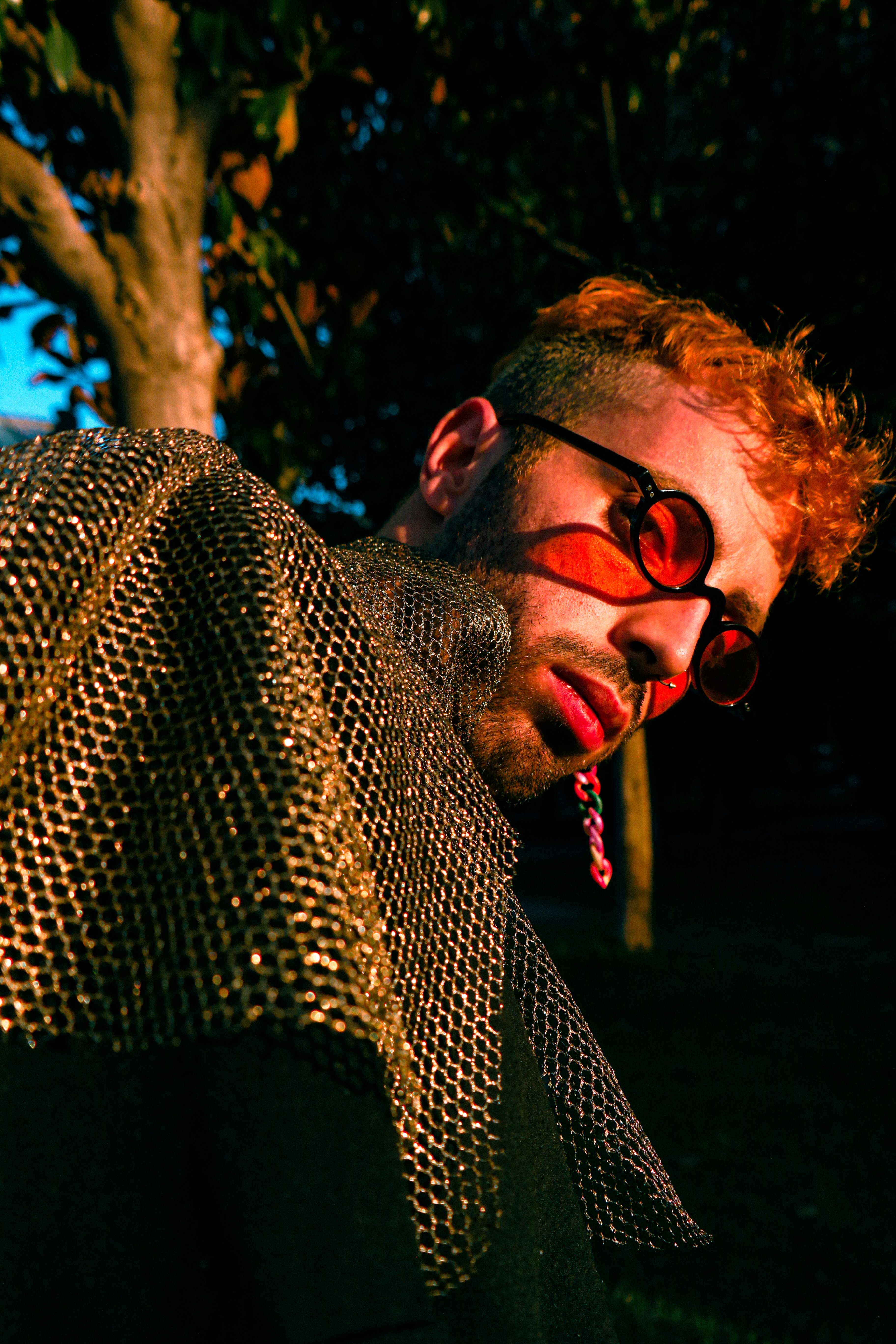 Fashionable man with red hair and sunglasses in a chainmail hood poses outdoors in dramatic lighting.