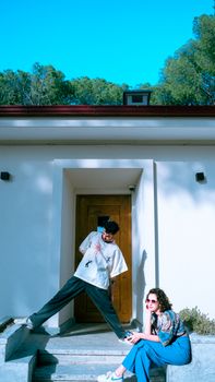 Two young people pose playfully in front of a modern house entrance with wooden doors.