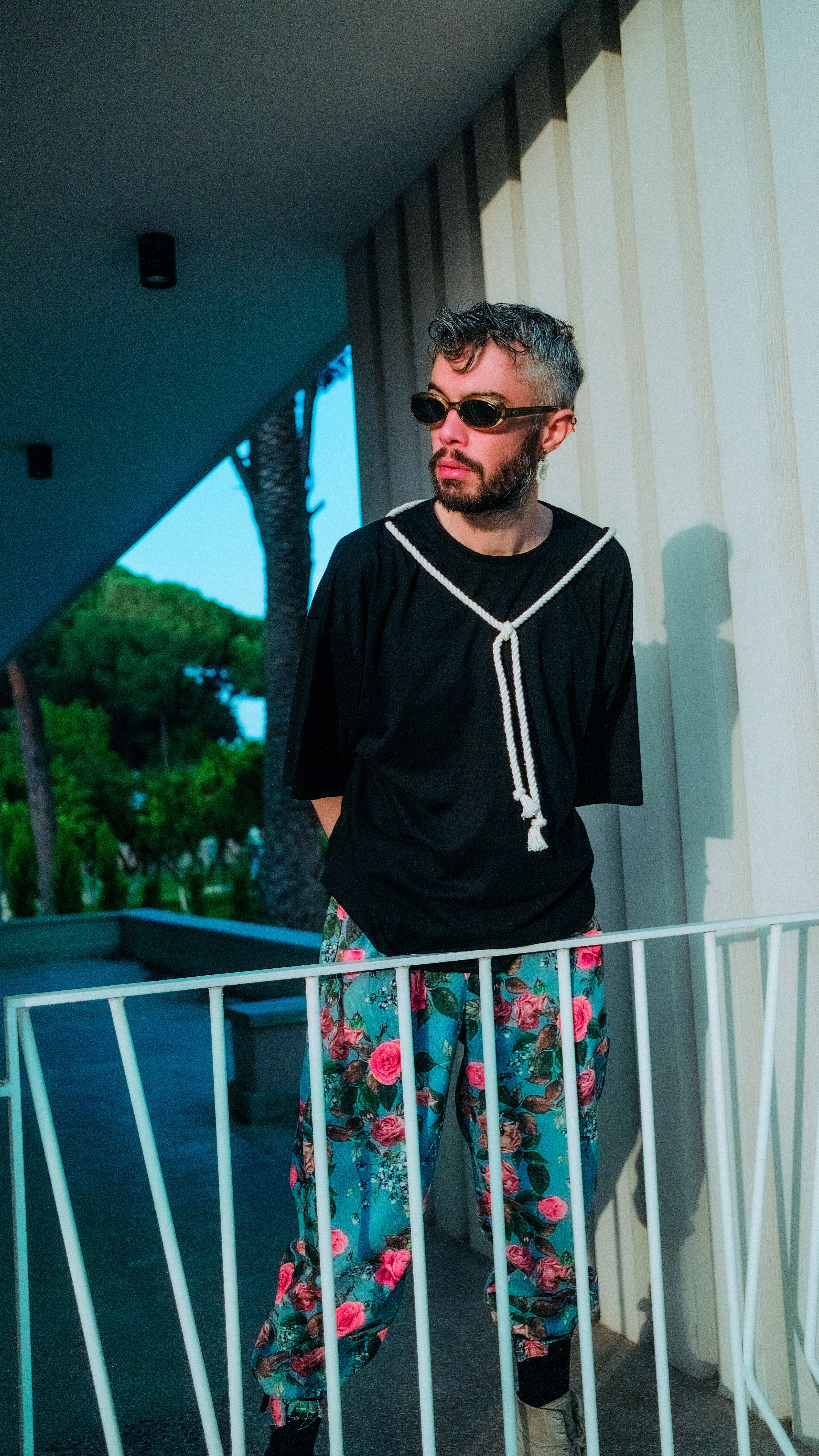 Homem fashion com óculos de sol posando do lado de fora com calças florais e camiseta preta.