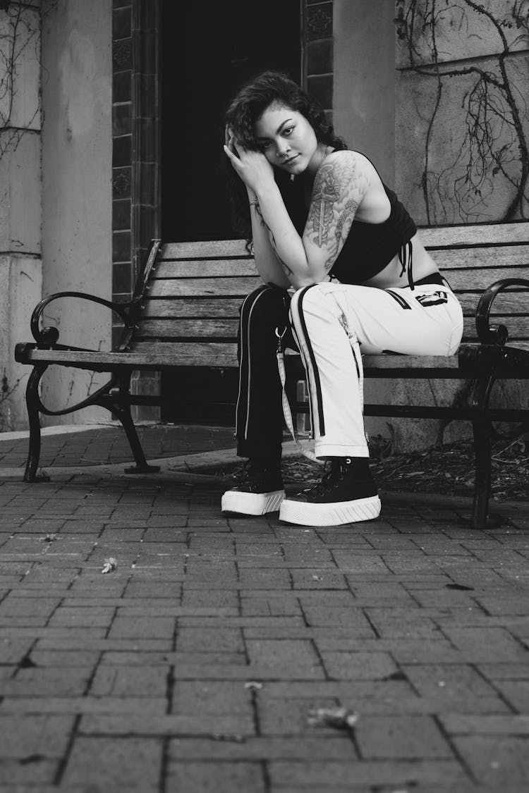 Young Woman In Black And White Pants, Crop Top And Platform Sneakers Sitting On A Bench