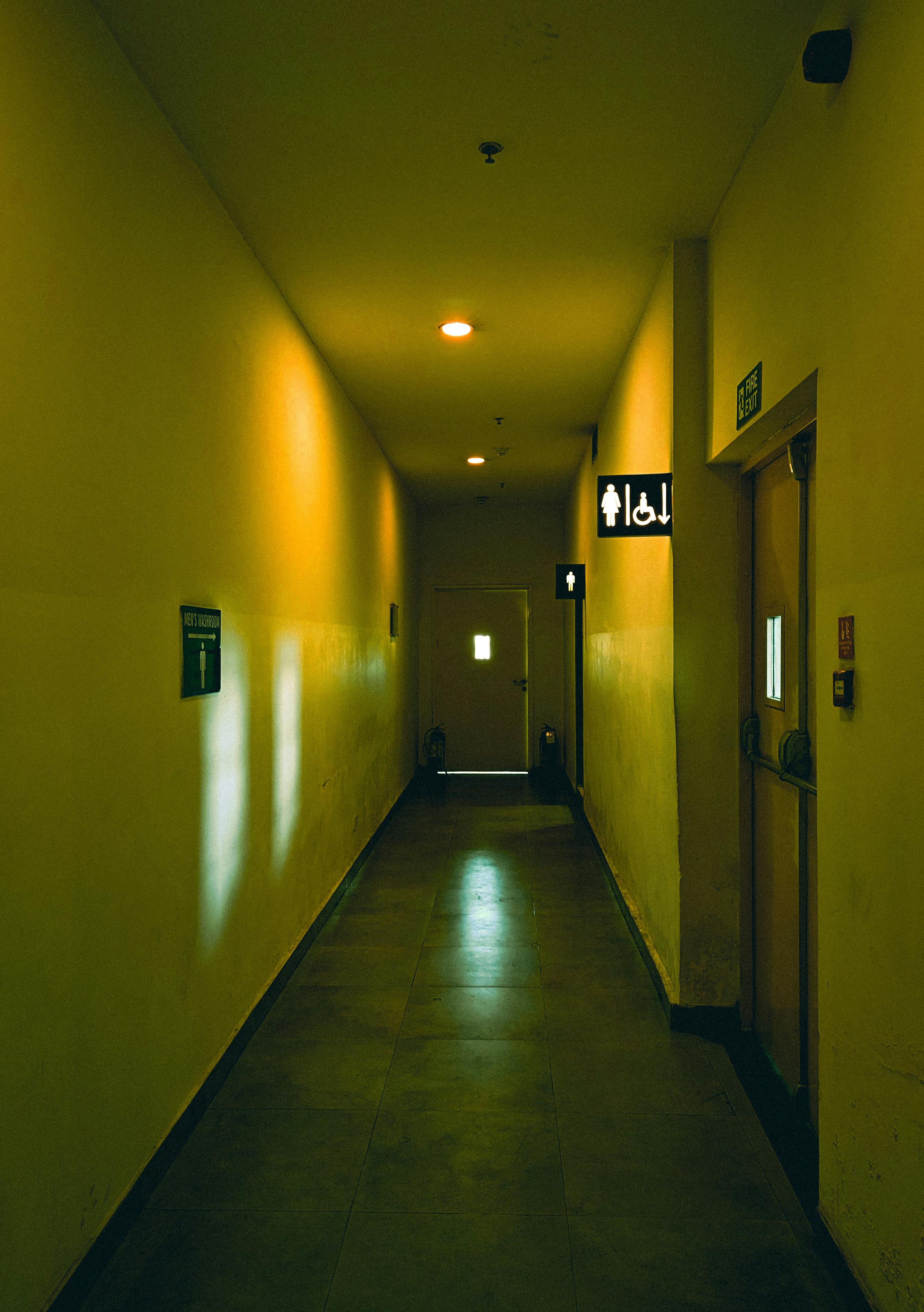 Free An empty corridor with yellow walls and ceiling lights, featuring restroom signs. Stock Photo