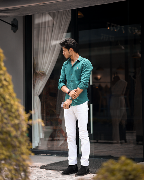 Free A fashionable young man in a green shirt and white pants standing by a glass door in an urban environment. Stock Photo