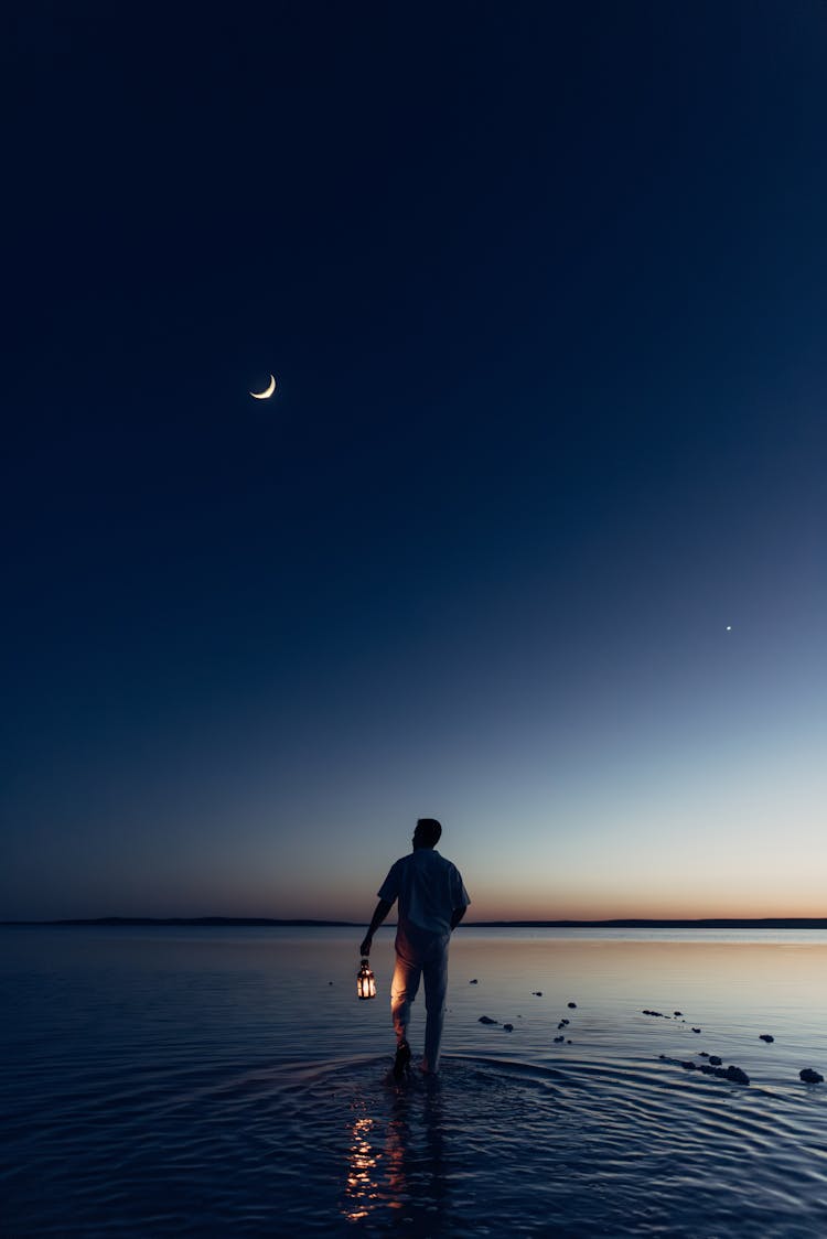 Crescent On Clear Sky Over Man Walking With Lanter On Shore At Night