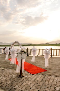 Beautiful outdoor wedding setup with red carpet and floral decorations by a tranquil lakeside at sunset.