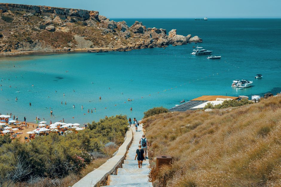 Scenic view of a beach in Malta with people, blue sea, and yachts in the summer sun.