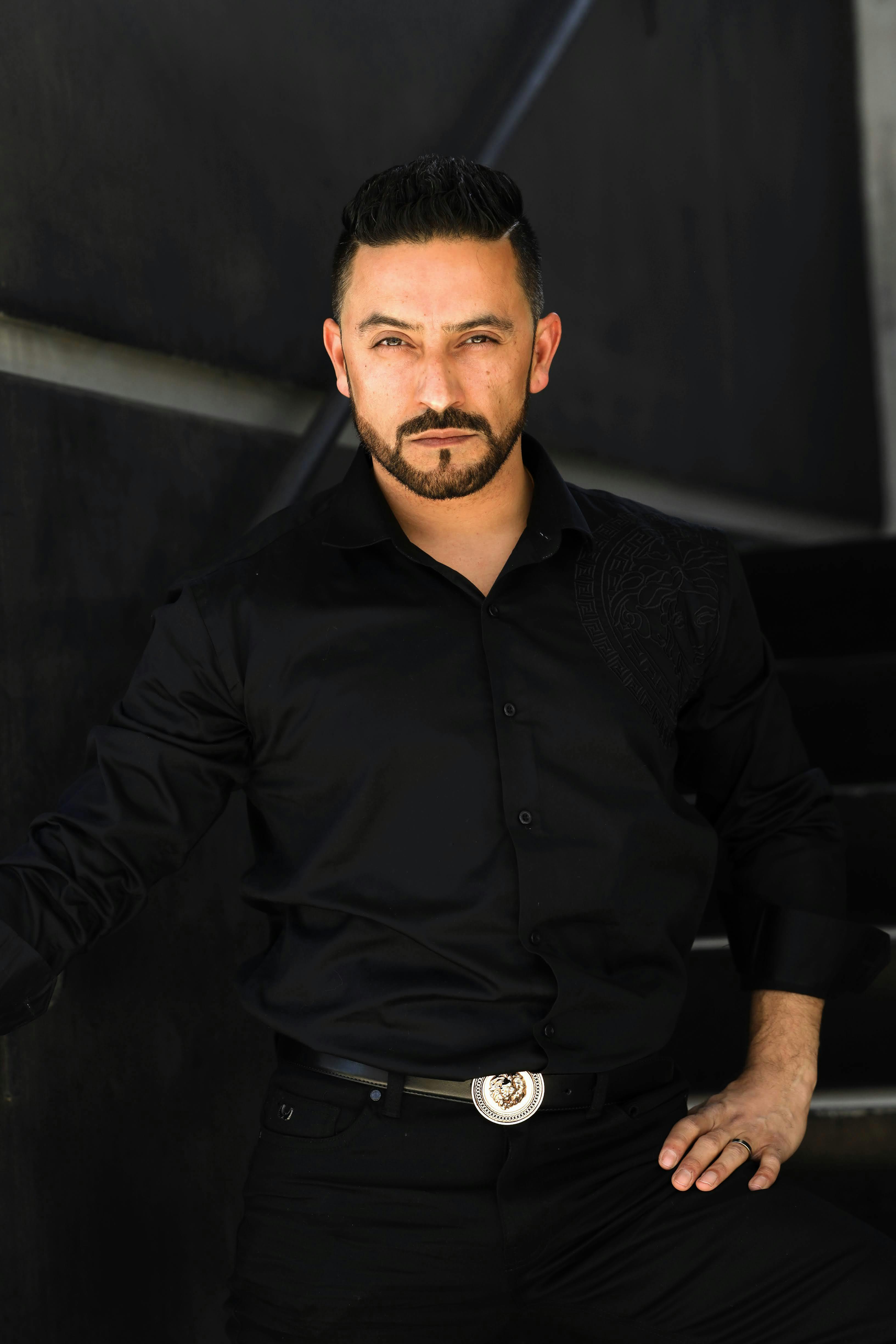 A confident man with a beard poses stylishly in black attire outdoors in Los Angeles.