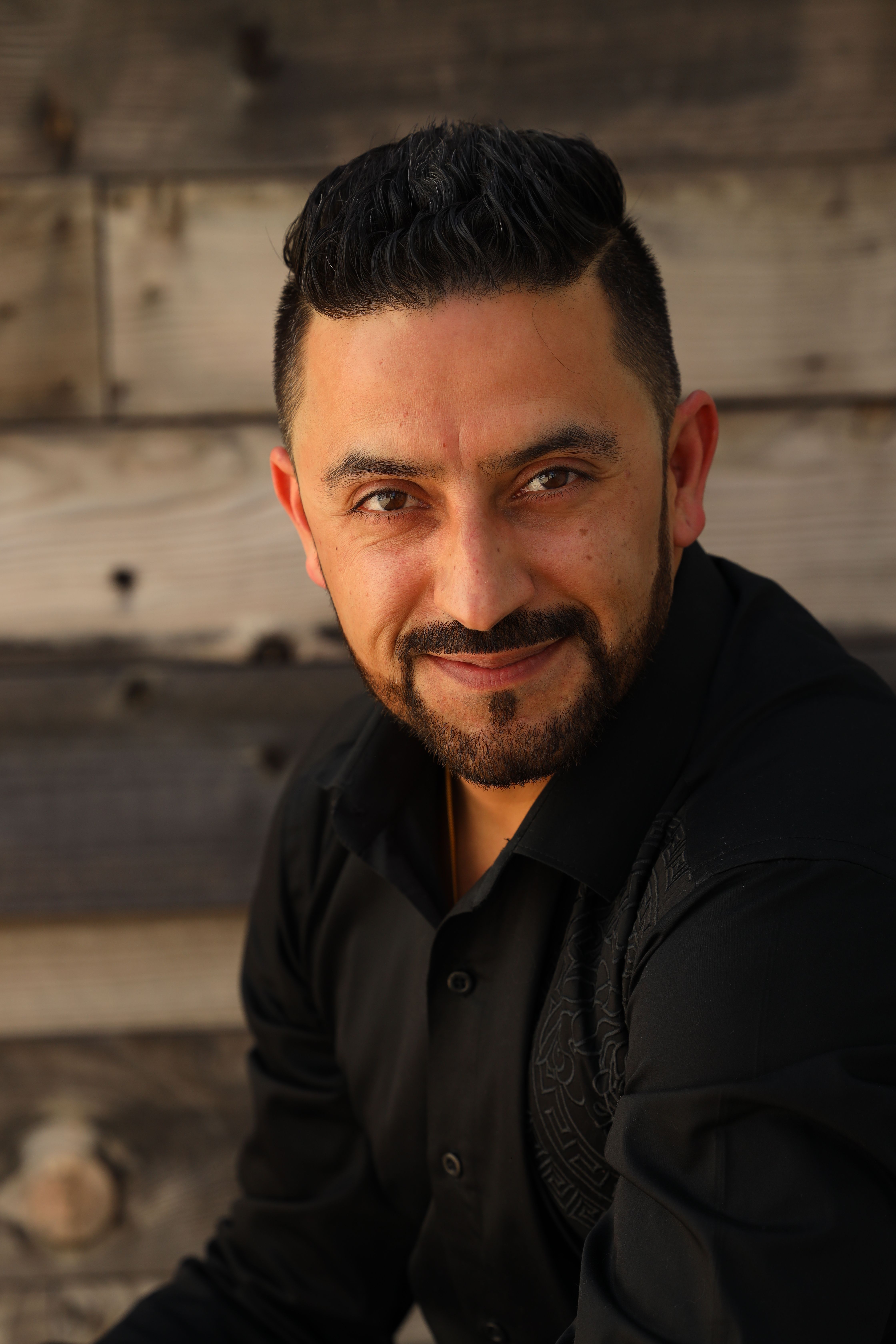 Smiling man with black hair and beard posing outdoors against wooden background.