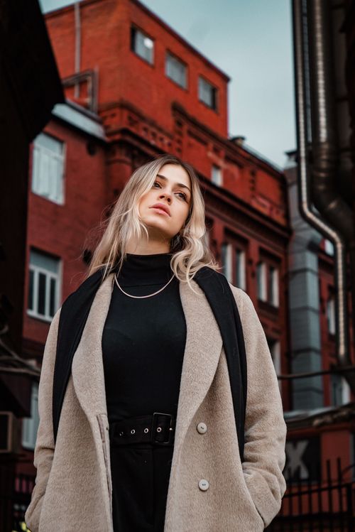 Free Fashionable woman in a beige coat standing confidently in a cityscape with red brick buildings. Stock Photo