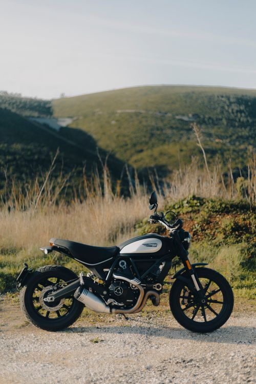 Free A Ducati Scrambler motorcycle parked on a dirt road with rolling hills in the background. Stock Photo