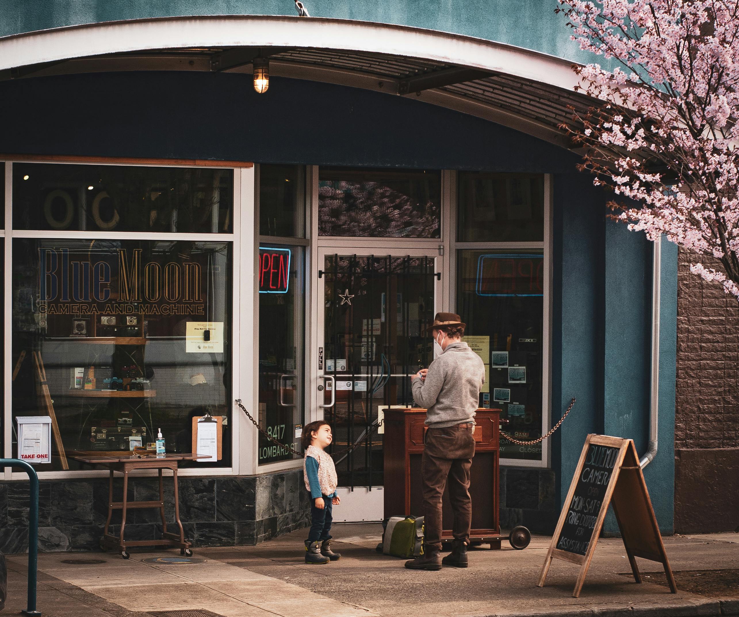 A man and child interact in front of Blue Moon Camera shop, exuding urban charm.