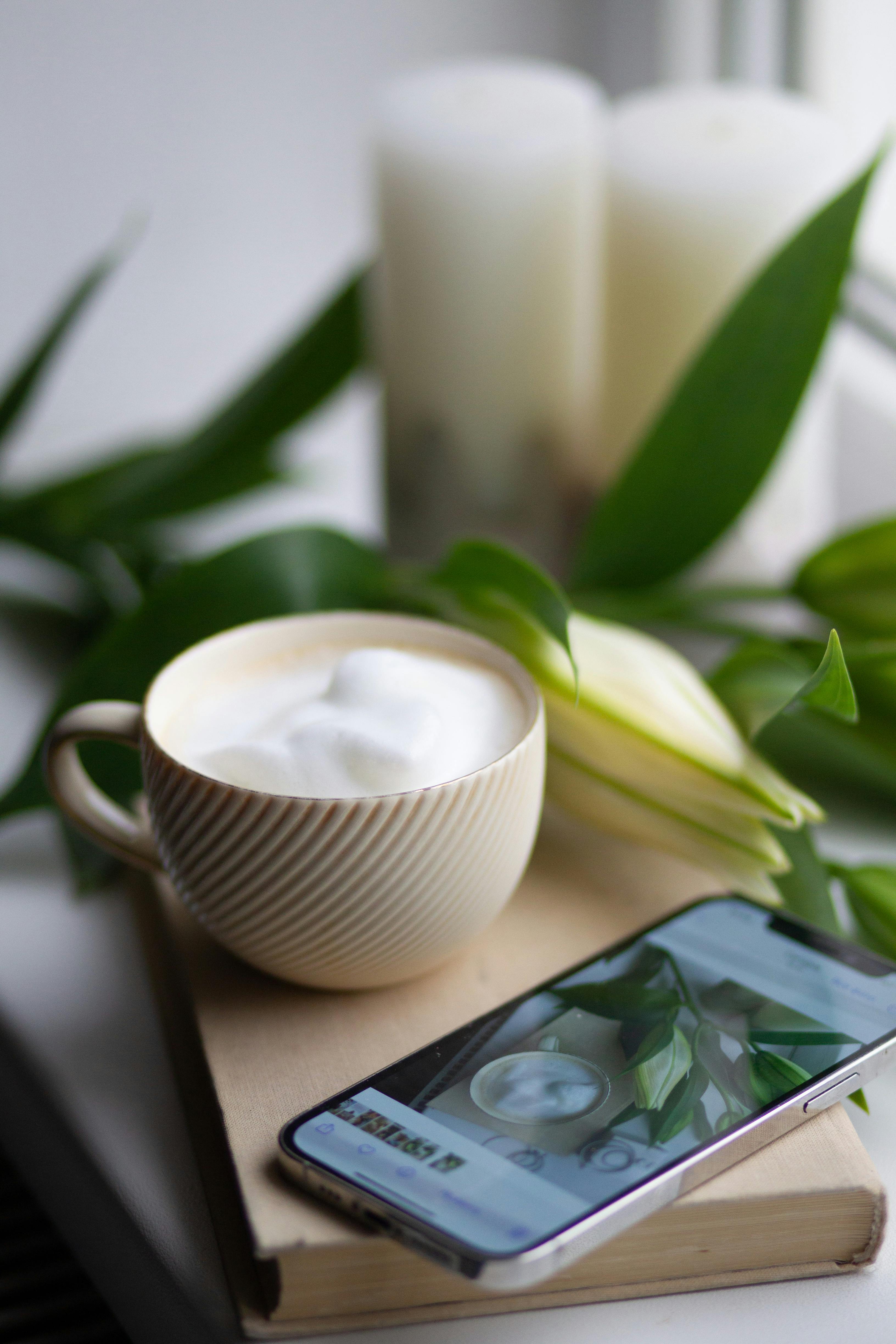 A warm cup of coffee with whipped cream sits on a book next to a smartphone and an Easter lily.