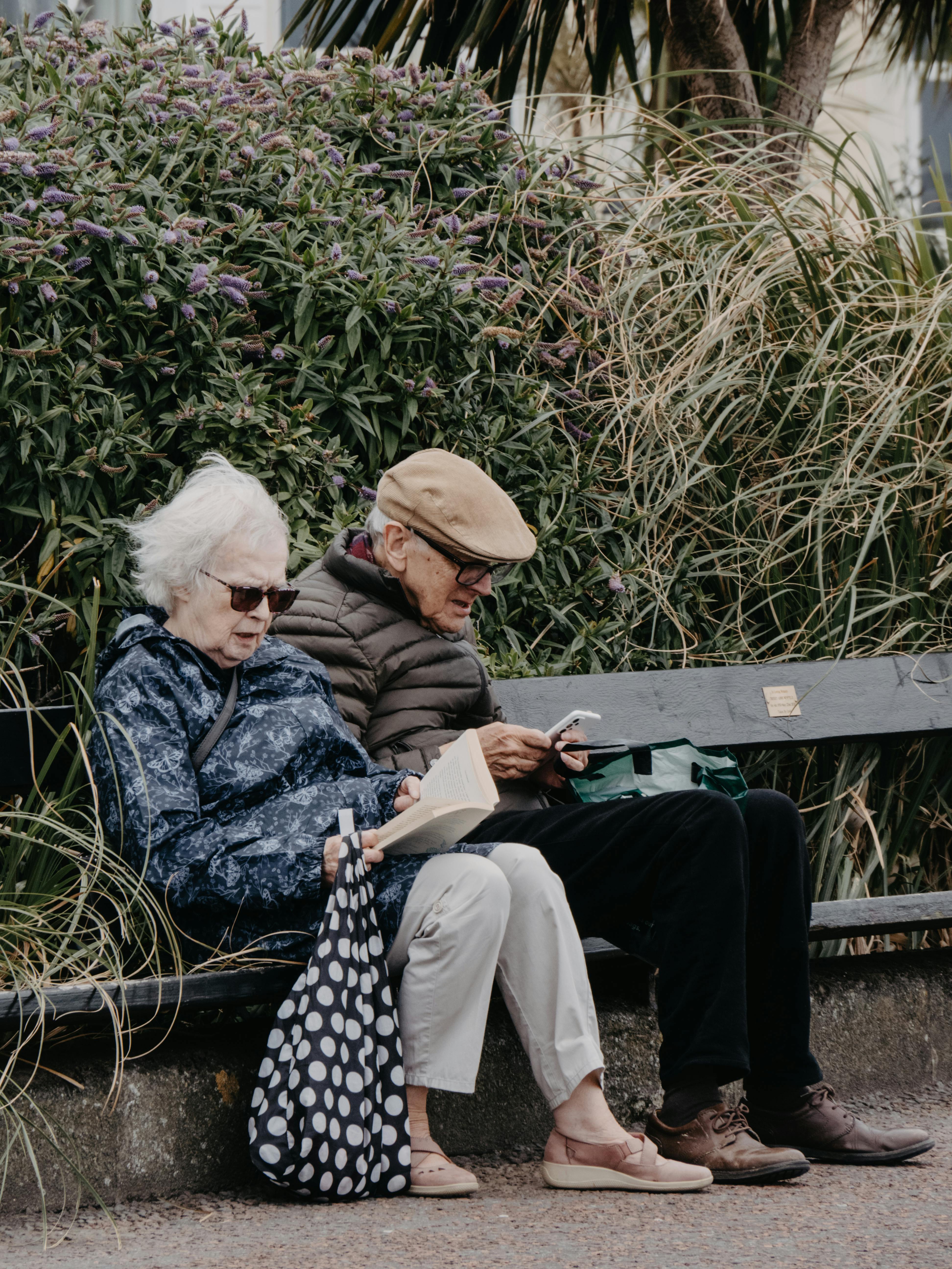 Senior couple enjoying a quiet moment reading on a park bench surrounded by greenery.