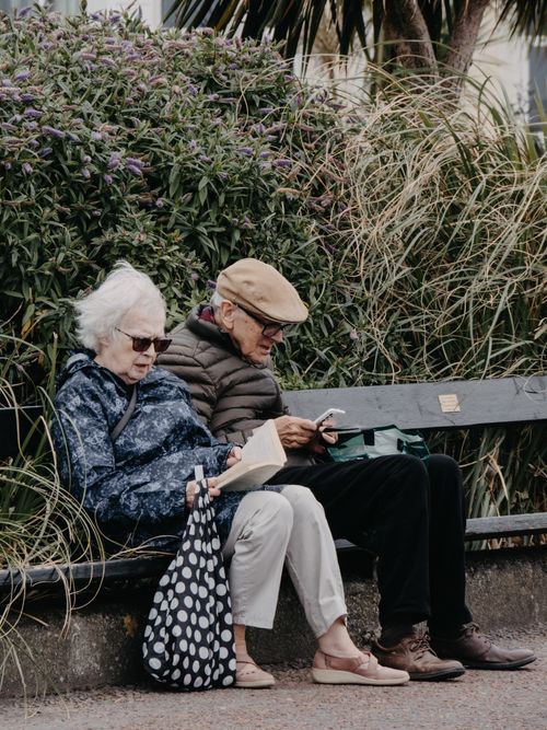 Free Senior couple enjoying a quiet moment reading on a park bench surrounded by greenery. Stock Photo