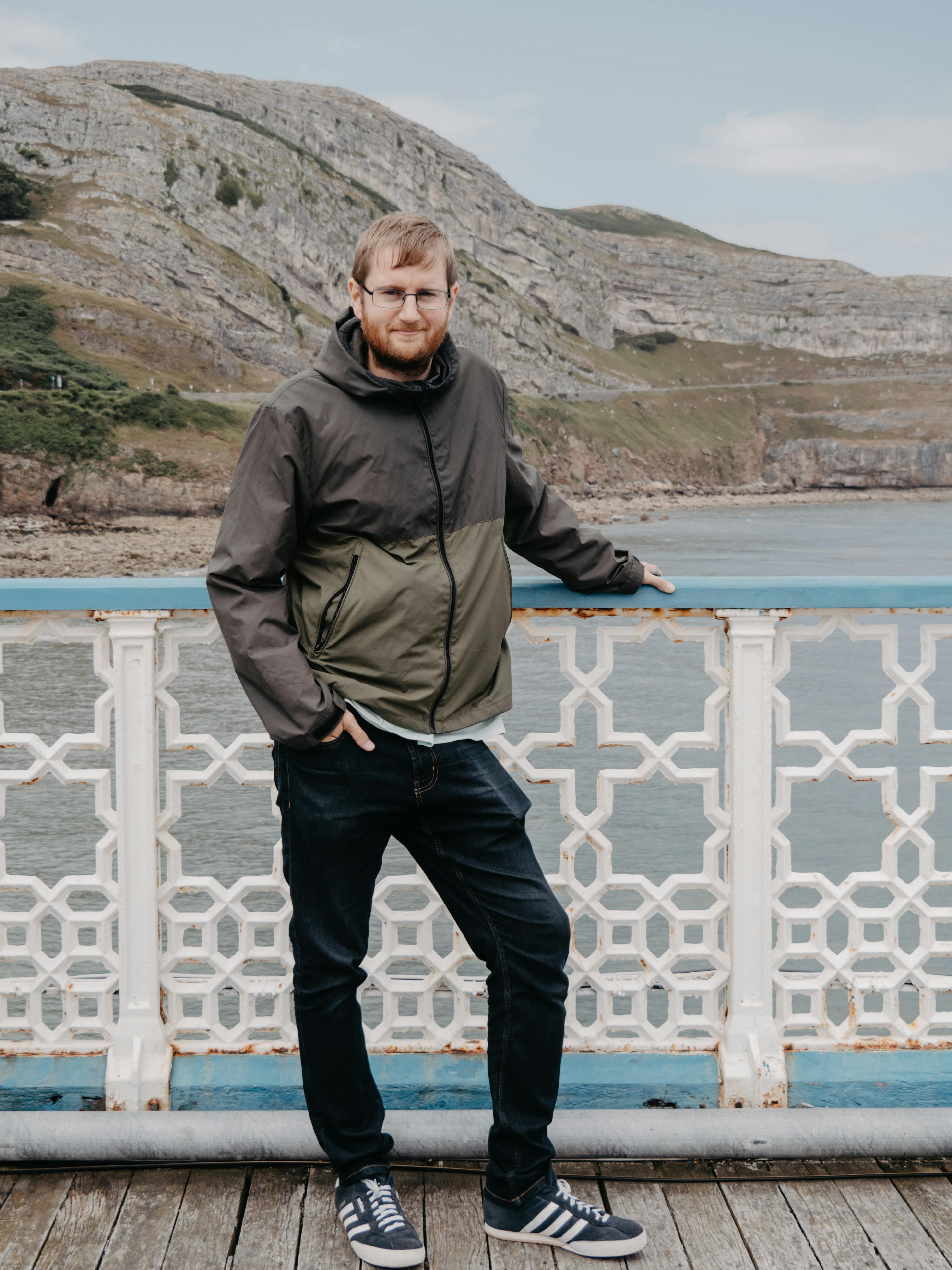 Man standing on Llandudno Pier, Wales, against a rocky hillside backdrop.