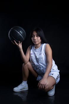 Young woman basketball player crouching, holding a ball against a dark background.