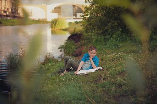 Free A thoughtful boy reading a book on a grassy riverbank during a sunny summer day. Stock Photo