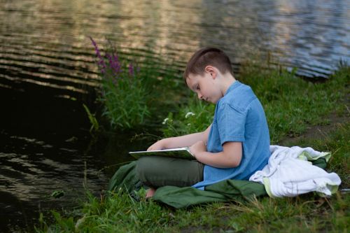 Free Boy reading by the riverbank, enjoying summer leisure outdoors. Stock Photo