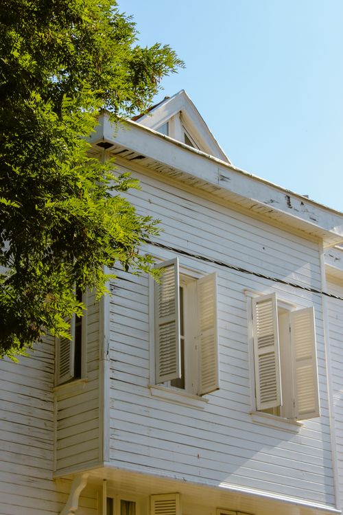 Free Charming white house facade with open shutters capturing sunlight beside lush green foliage. Stock Photo