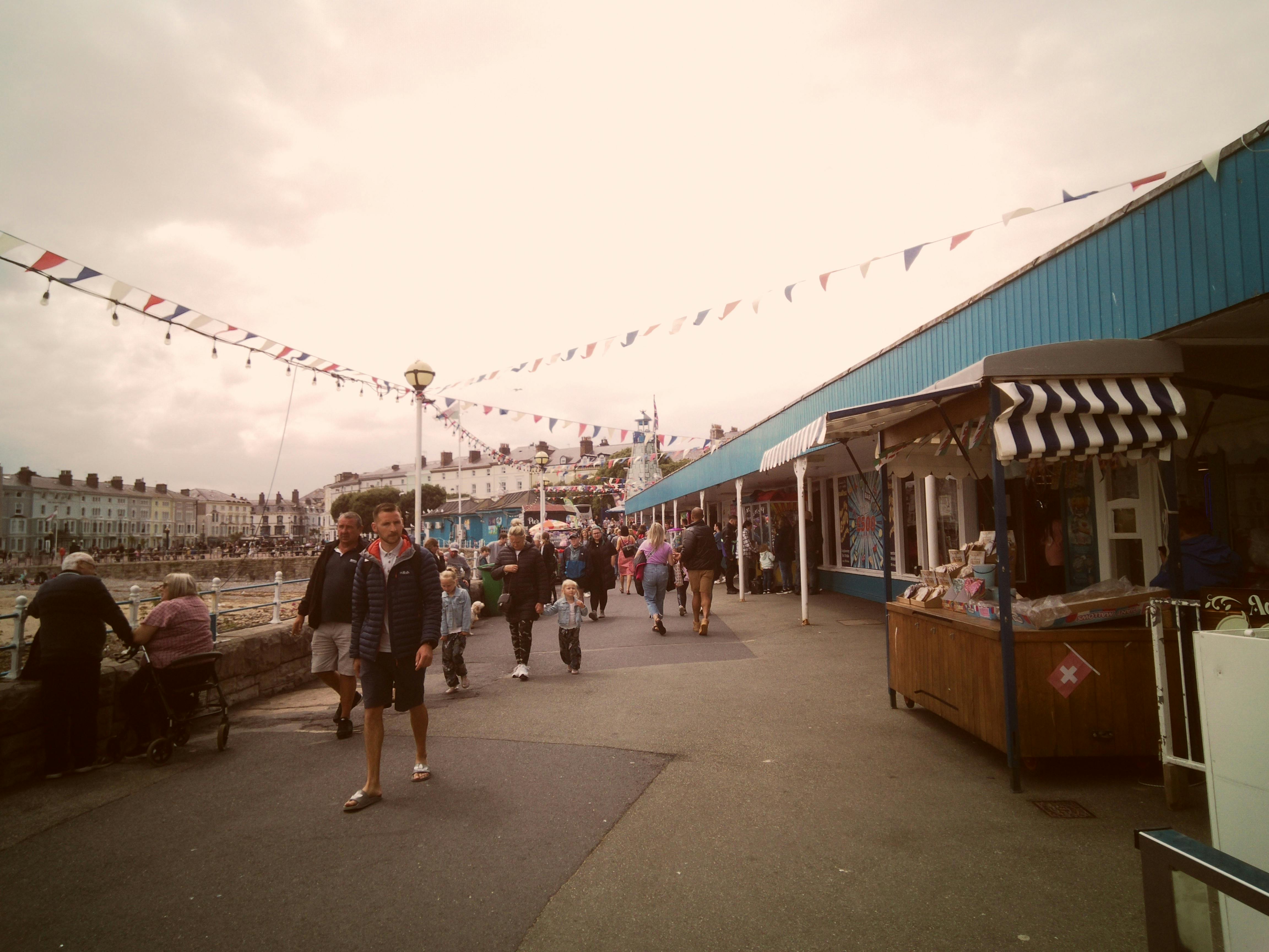 Lively outdoor market in Llandudno, Wales with people, shops, and festive flags.